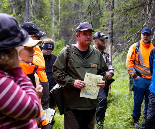 Kari-Pekka Poutanen metsäalan opiskelijoidenkanssa metsässä kädessään paperi.