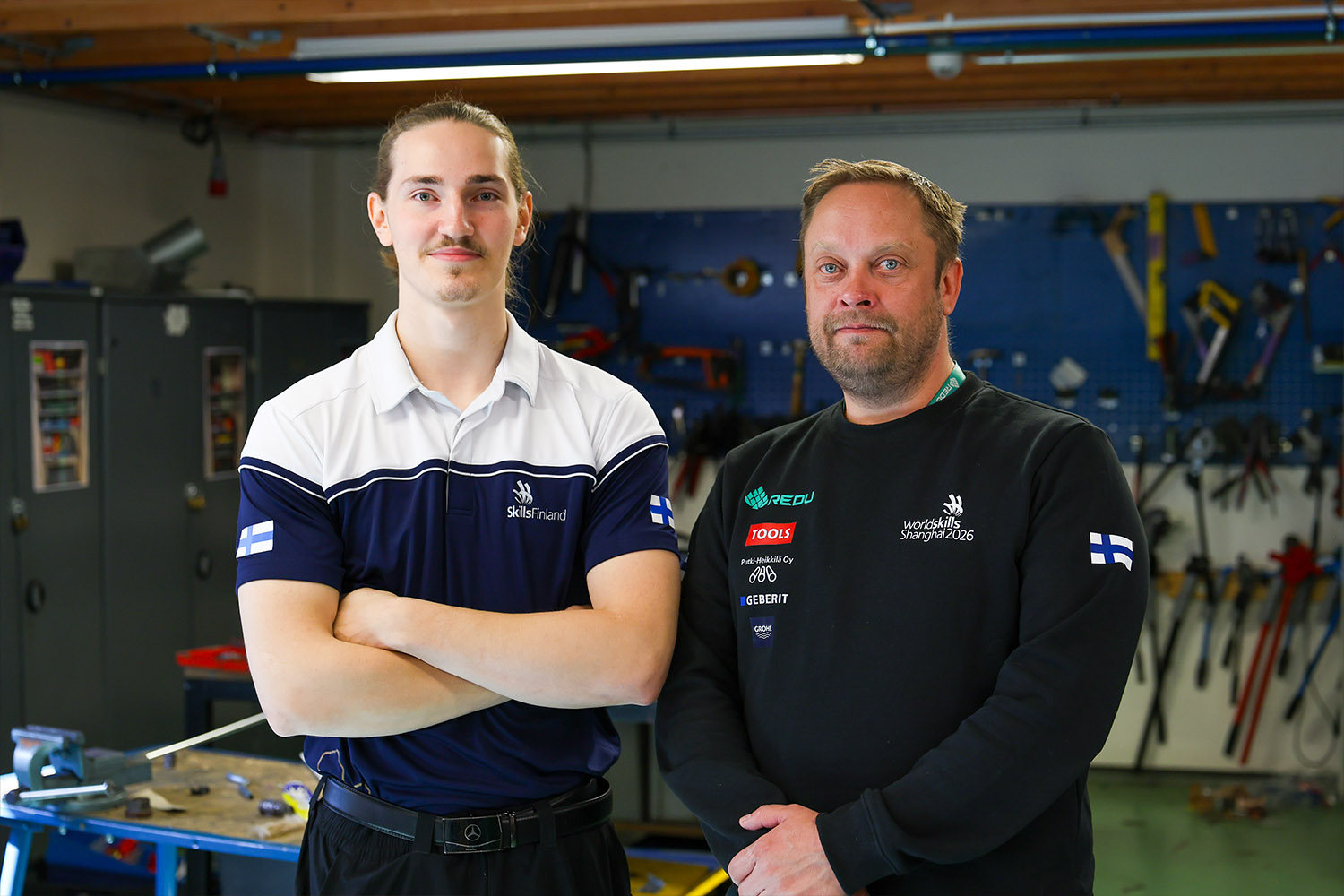 WorldSkills-kilpailija ja valmentaja seisovat talotekniikan opetustilassa työkaluseinän edessä.