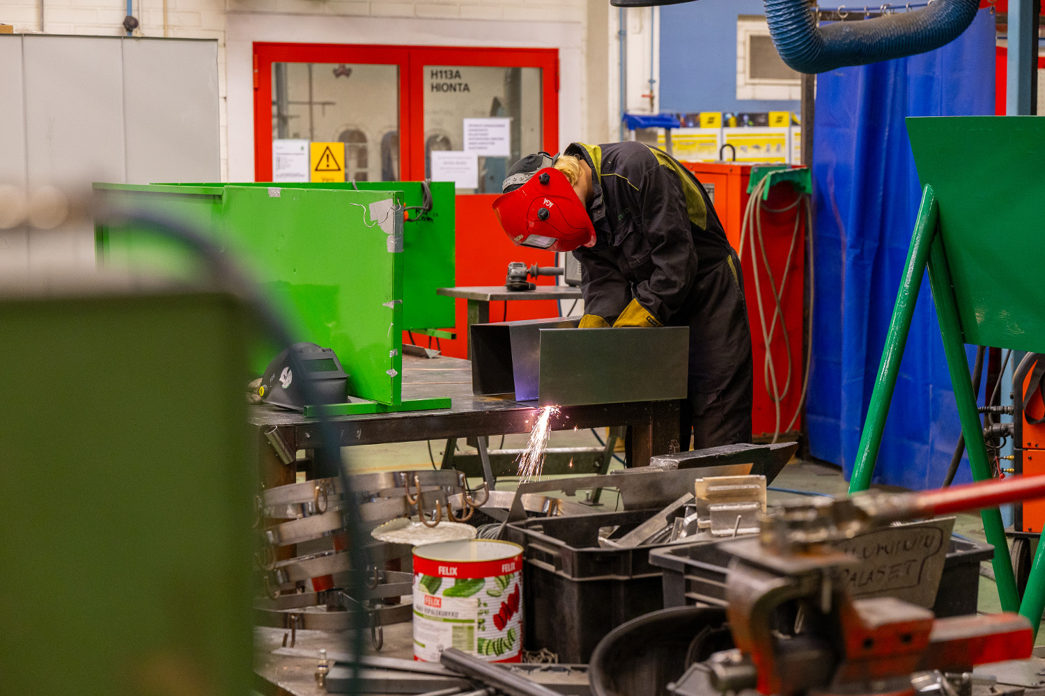 Henkilö käsittelemässä suojavarusteissa rälläkällä metallia, josta tulee kipinöitä.