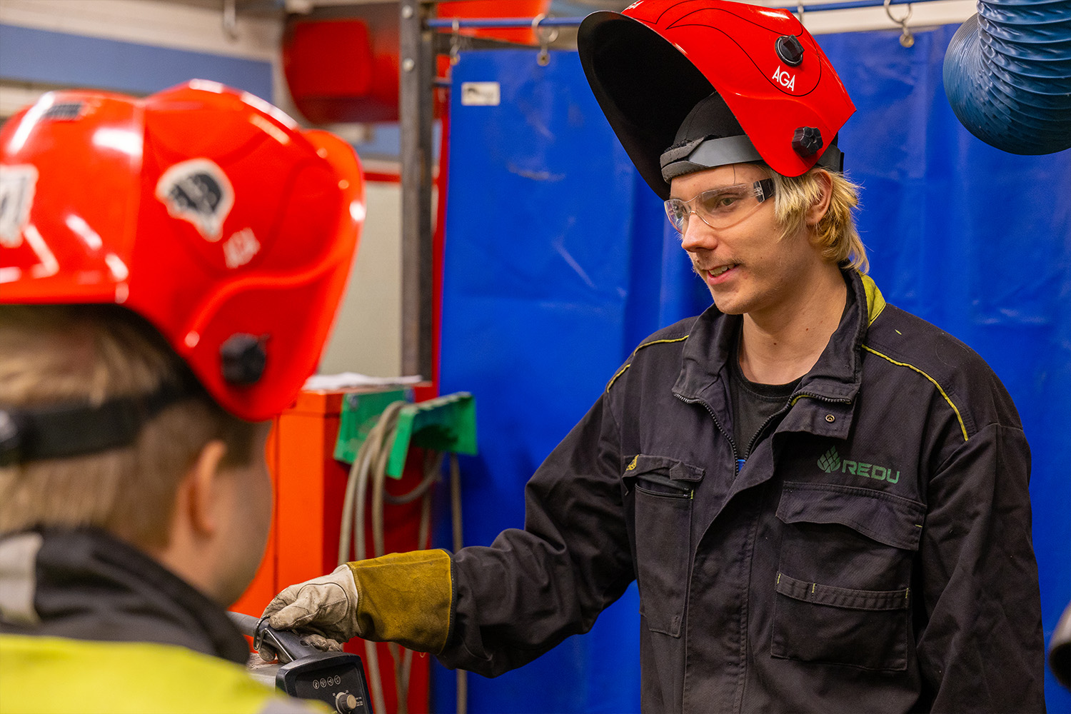 Henkilö hitsausmaski päässä hymyilemässä ja juttelemassa selin kameraanpäin olevalle henkilölle.